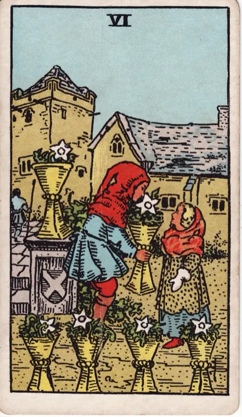 Six of Cups, from the 1909 Rider-Waite-Smith deck: a boy hands a girl a cup filled with white flowers in an old village courtyard, more flower-filled cups around them.
