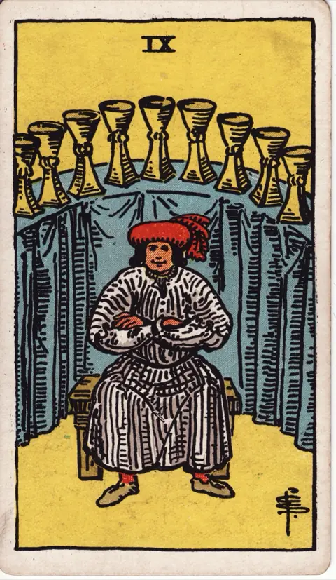 Nine of Cups, from the 1909 Rider-Waite-Smith deck: a contented man sits with folded arms in front of a curved table draped in blue, nine cups lined up on it behind him.