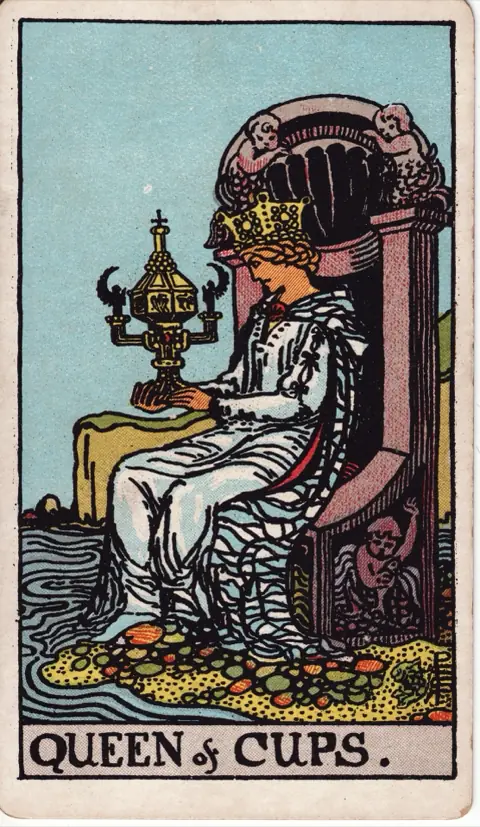 Queen of Cups, from the 1909 Rider-Waite-Smith deck: a queen sits on a throne at the edge of the sea, gazing at an ornate covered cup with angel handles that she holds in both hands.