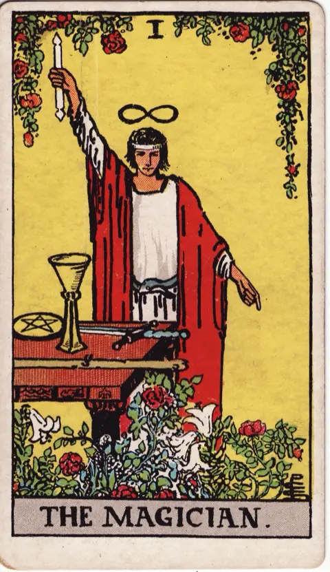 The Magician, from the 1909 Rider-Waite-Smith deck: a magician in a red robe raises a wand to the sky and points to the ground, an infinity sign over his head, with a cup, a sword, a pentacle and a wand on the table before him among roses and lilies.