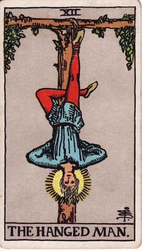 The Hanged Man, from the 1909 Rider-Waite-Smith deck: a man hangs upside down by one foot from a living T-shaped tree, his other leg bent behind his knee, his hands behind his back and a halo around his calm face.
