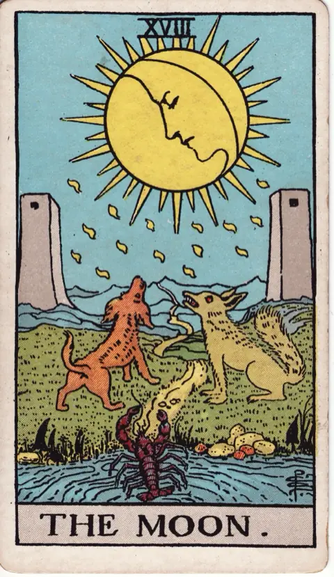 The Moon, from the 1909 Rider-Waite-Smith deck: a moon with a face shines between two towers, a dog and a wolf howl up at it, and a crayfish climbs out of a pool onto a path that winds into the hills.