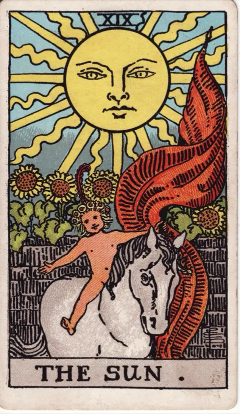 The Sun, from the 1909 Rider-Waite-Smith deck: a naked child with a red feather in his hair rides a white horse under a large smiling sun, holding a red banner, sunflowers growing over the wall behind.