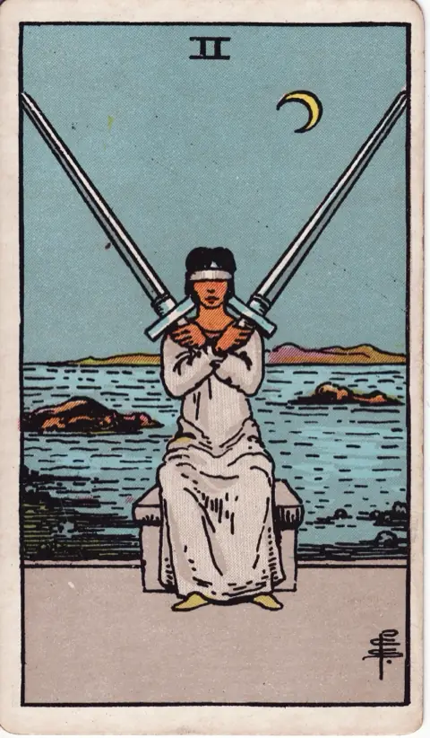 Two of Swords, from the 1909 Rider-Waite-Smith deck: a blindfolded woman in white sits on a stone bench holding two crossed swords over her chest, the sea and a crescent moon behind her.