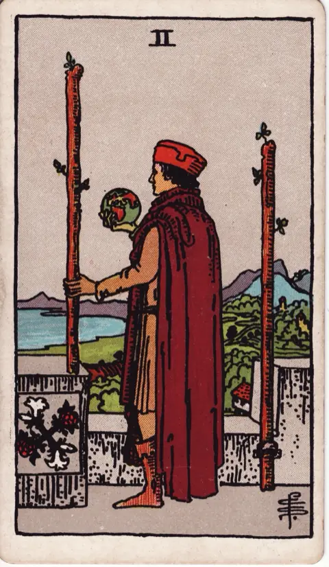 Two of Wands, from the 1909 Rider-Waite-Smith deck: a man in a red cap stands on castle battlements holding a small globe and one wand, a second wand fixed to the wall, looking out over sea and land.