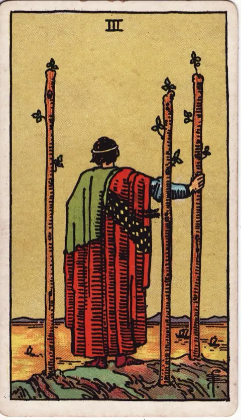 Three of Wands, from the 1909 Rider-Waite-Smith deck: a man in a red and green robe stands on a cliff with his back to us beside three planted wands, watching ships cross a golden sea.