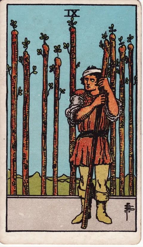 Nine of Wands, from the 1909 Rider-Waite-Smith deck: a man with a bandaged head leans on a wand and looks warily over his shoulder, eight wands standing in a row behind him.